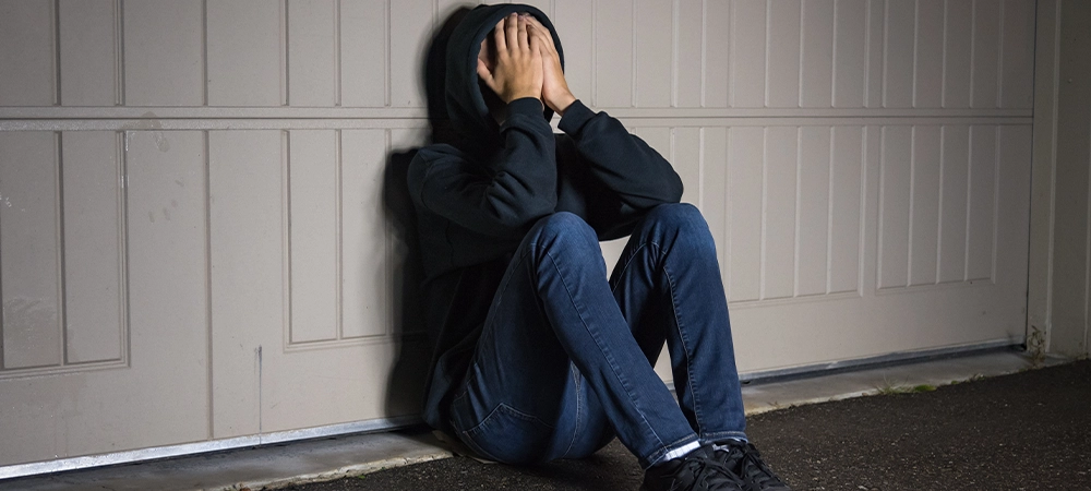 depressed male teenager sitting on ground in front of garage door and covering his face